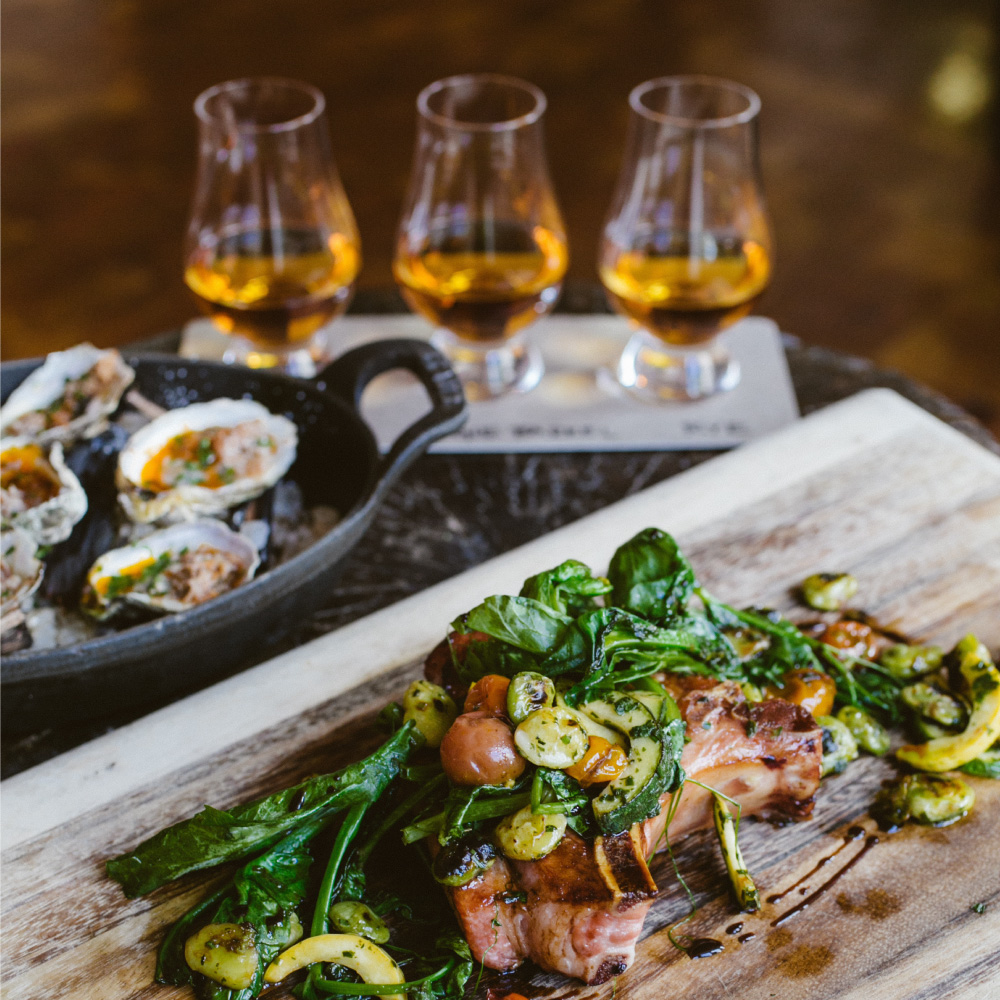 Grilled Meat and vegetables on a wooden tray. Background 3 bourbon dram glasses and seafood plate